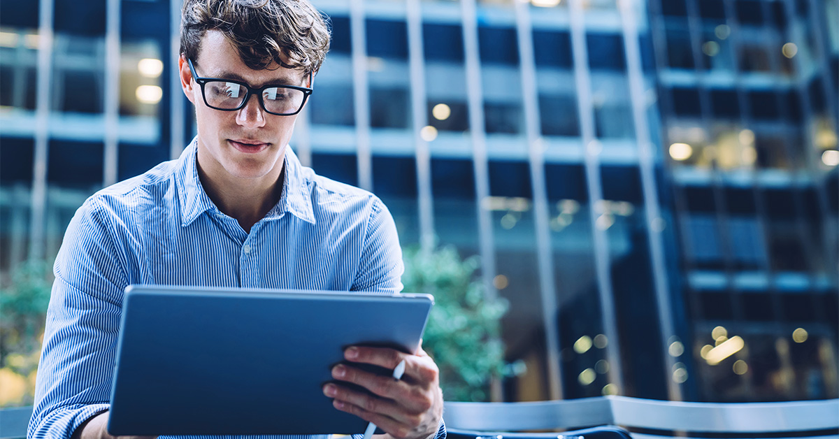 Homem branco usando óculos e uma camisa azul listrada sentado na frente de um prédio espelhado segurando um tablet cinza.