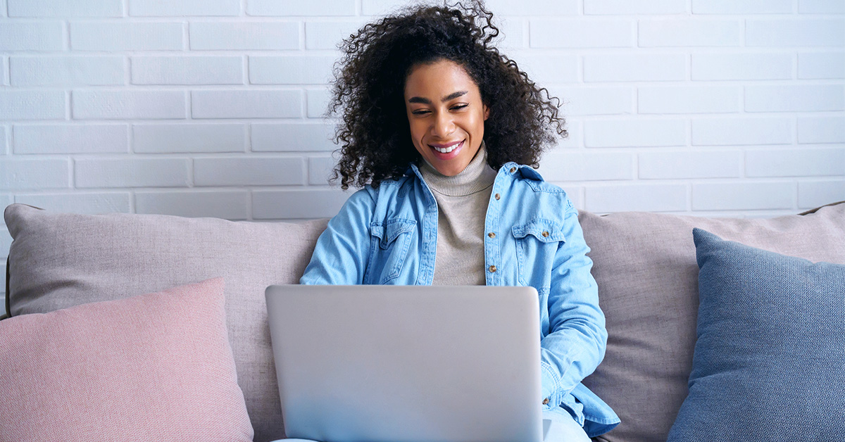 Uma mulher negra, de cabelos longos e crespos, vestida com uma jaqueta jeans azul, está sentada no sofá, pesquisando informações no notebook.
