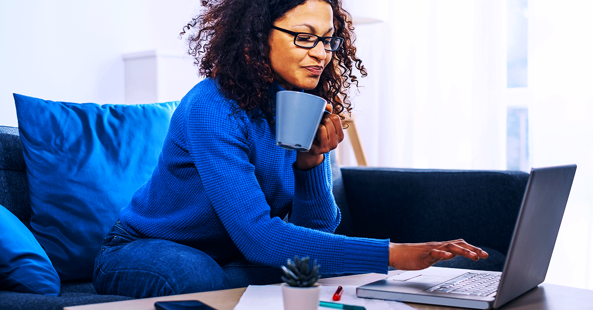 Mulher negra vestindo blusa de frio azul e calça jeans sentada em um sofá cinza escuro mexendo em um notebook cinza claro.
