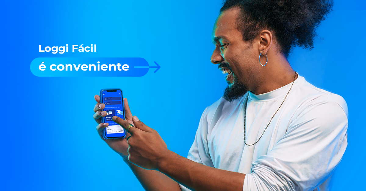 Um homem preto, com o cabelo amarrado e vestindo uma blusa branca, sorri enquanto segura um celular que mostra a tela inicial do Loggi Fácil, o serviço de envios da Loggi.