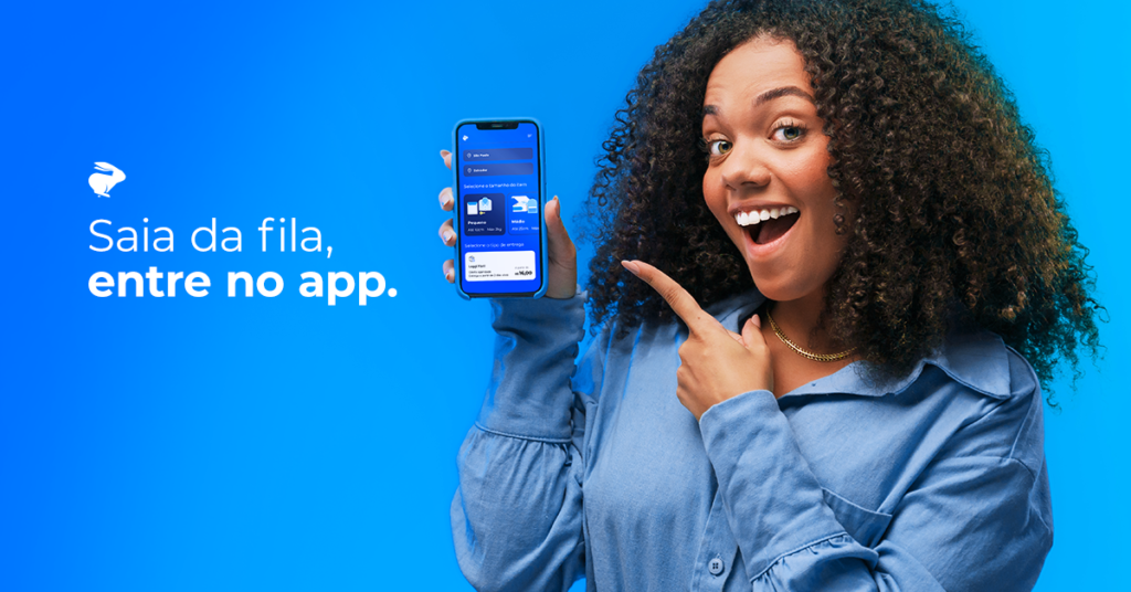 Uma muler negra, de cabelos encaracolados, sorri enquanto segura um celular que mostra a tela inicial do Loggi Fácil, o serviço da Loggi. 