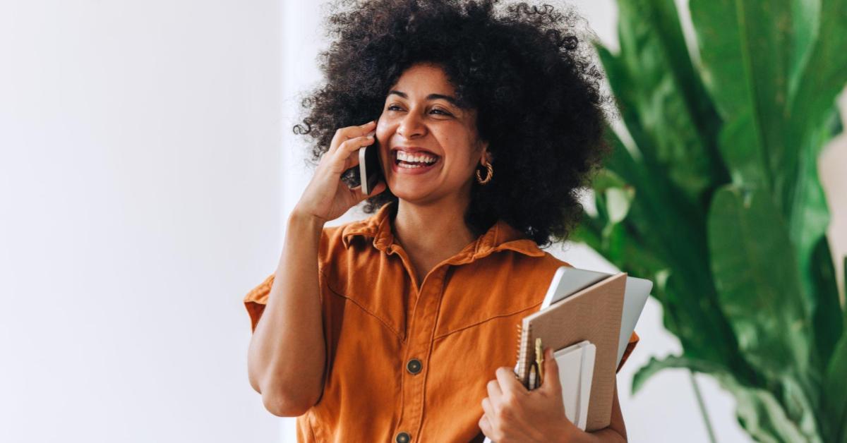 Uma mulher preta, com black power e um sorriso no rosto, fala ao celular enquanto segura alguns cadernos em uma das mãos.