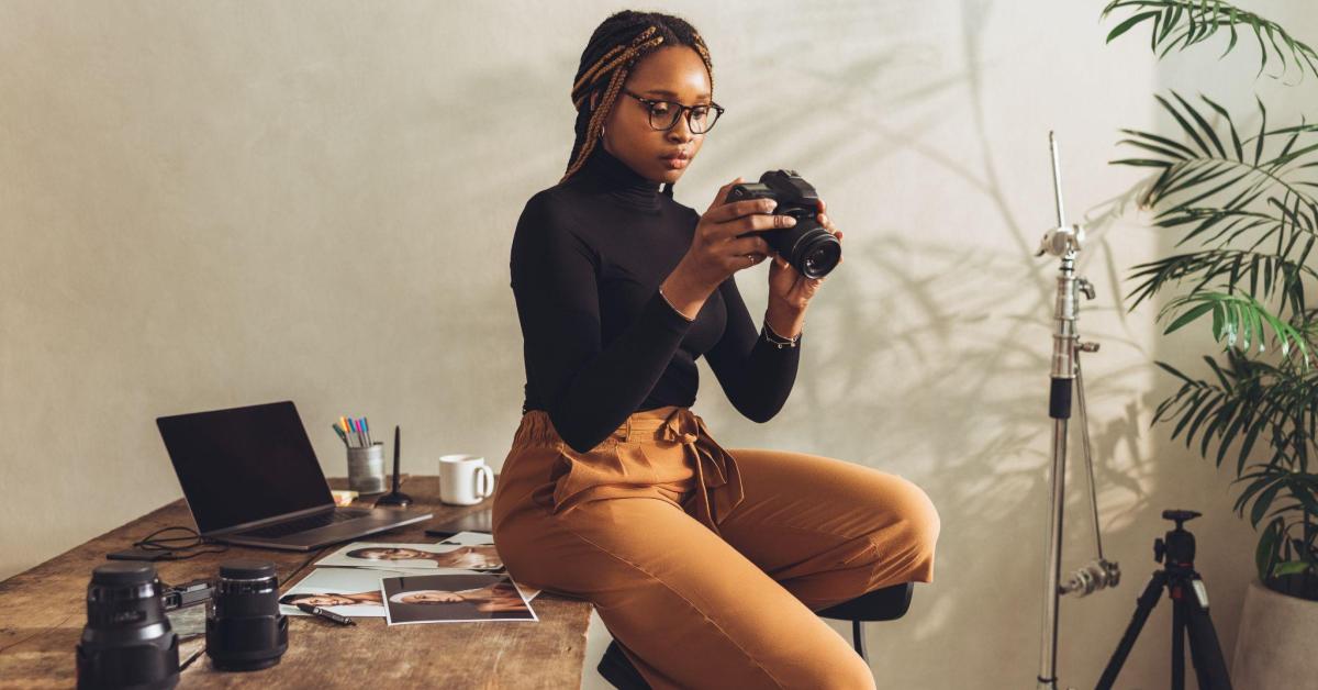 Uma mulher negra, com óculos e cabelos com dreads, veste uma blusa preta e calça marrom. Sentada numa mesa de madeira com diversas fotografias de modelos, um notebook e caneta, ela observa na câmera que segura, a foto que tirou recentemente. 