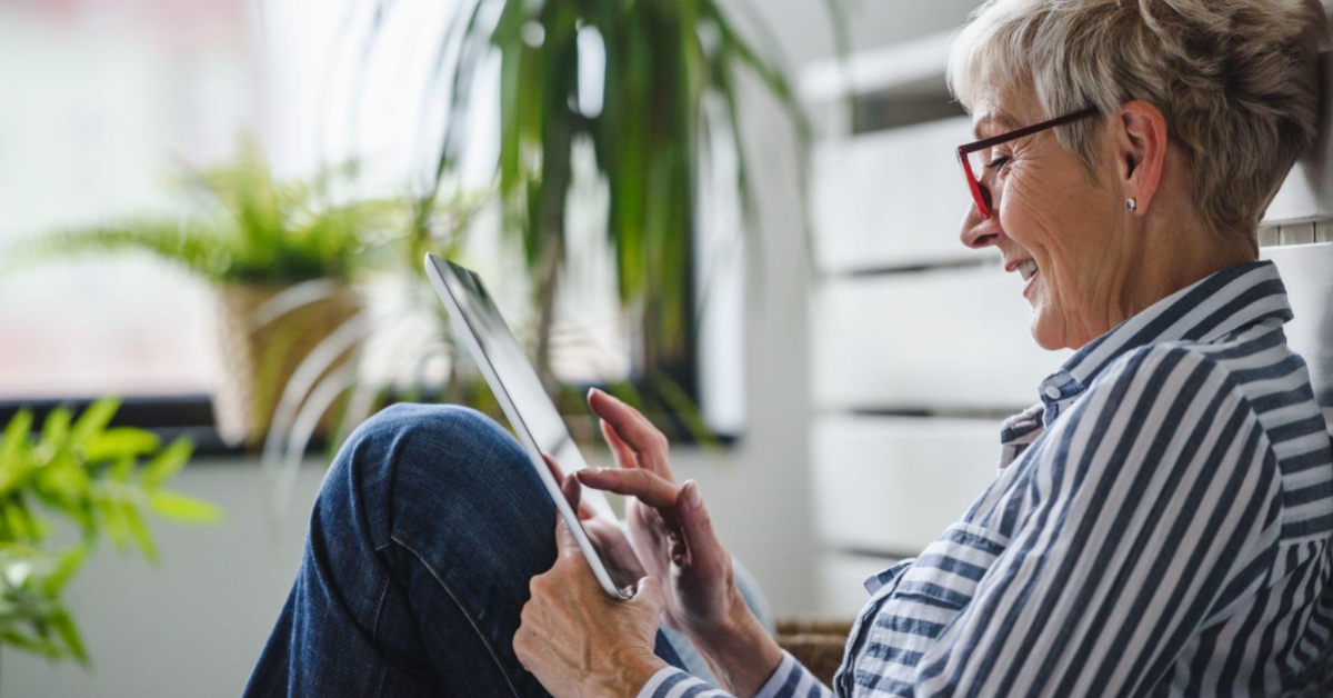 Uma mulher de cerca de 70 anos, de cabelos curtos grisalhos, está sentada no chão enquanto mexe em um tablet. Ela sorri para o aparelho, e veste óculos vermelhos, camisa listrada e calça jeans. Na imagem vemos a mulher de lado, enquanto ao fundo há diversas plantas compondo o cenário.