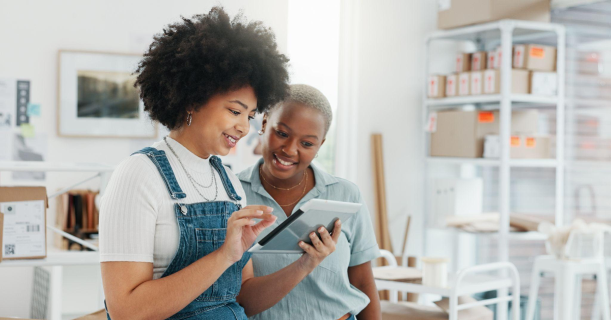 Duas mulheres ptreas, uma com black power, outra com o cabelo raspado e cinza, mexem em um tablet. Elas estão dentro de uma sala que remete a um espaço de estoque. A ideia da imagem é reforçar a organização necessária durante a Black Friday.
