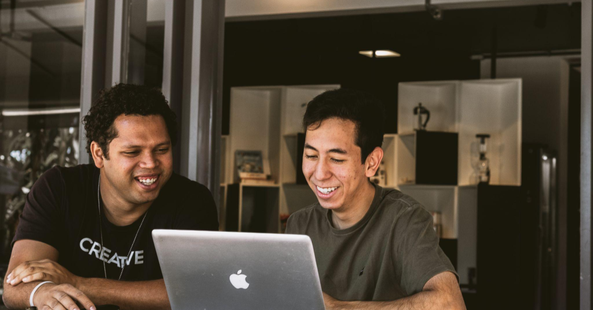 Dois homens estão sentados em um escritório, um ao lado do outro, olhando para a tela de um notebook. Um dos homens é preto, de cabelos curtos cacheados. Ele veste uma camiseta preta. O outro, é um homem amarelo, de cabelos pretos e curtos. Ele veste uma camisa verde-escura. Ambos sorriem para o que estão vendo na tela, fazendo uma associação ao momento em que consumidores leem avaliações no e-commerce sobre um produto que têm procurado.