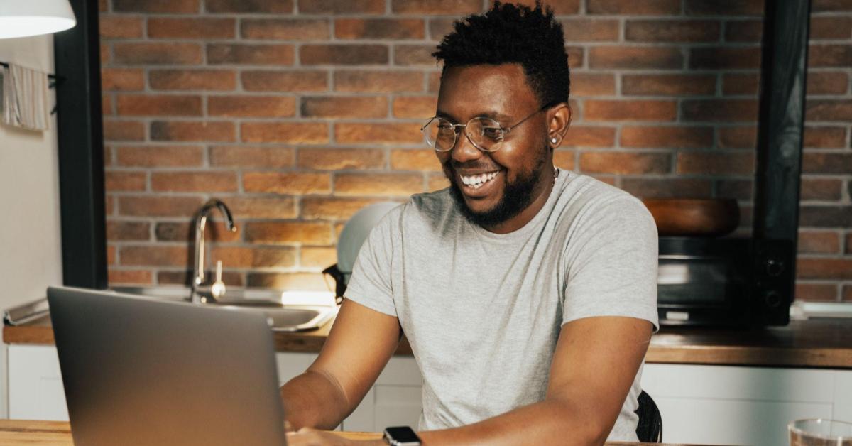Um homem preto, com dreads curtos, usa uma camiseta branca e óculos redondos de aros finos. Ele está sentado em uma sala, e sorri enquanto mexe em seu notebook. Uma alusão ao momento em que as pessoas buscam por suas intenções de consumo na black friday.