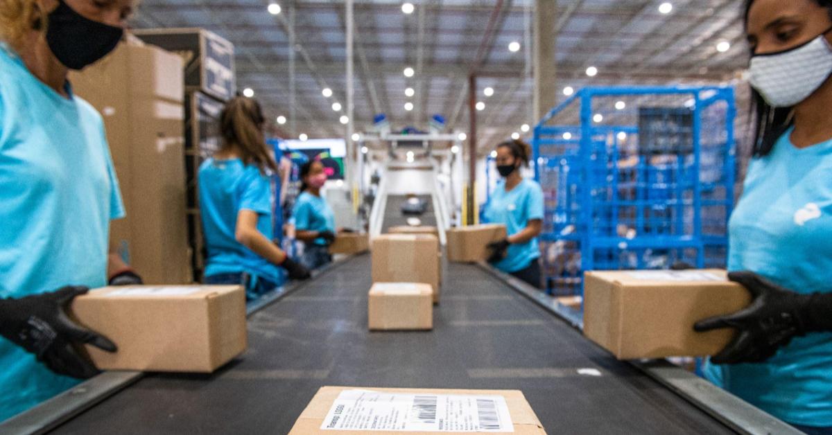 A imagem mostra o interior de um dos crossdockings da Loggi. A visão que a pessoa tem, é como se ela estivesse olhando o sorter, rampa em que os pacotes passam, de dentro. Na imagem, diversos pacotes passam pelos sortes, enquanto pessoas bipam as etiquetas. Esse é um dos processos necessários para o envio de encomendas.