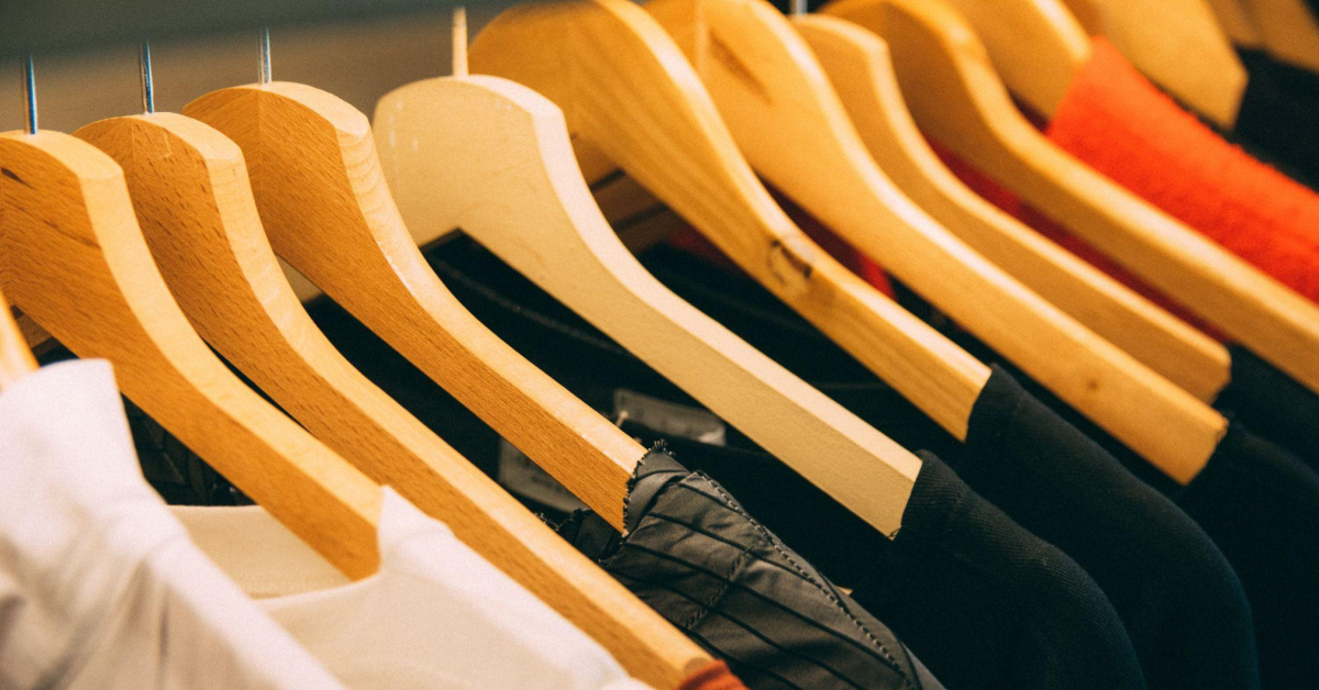 A imagem dá foco em uma arara de camisetas, penduradas em cabides de madeira. Esse é um possível produto para quem almeja ter um brechó online.
