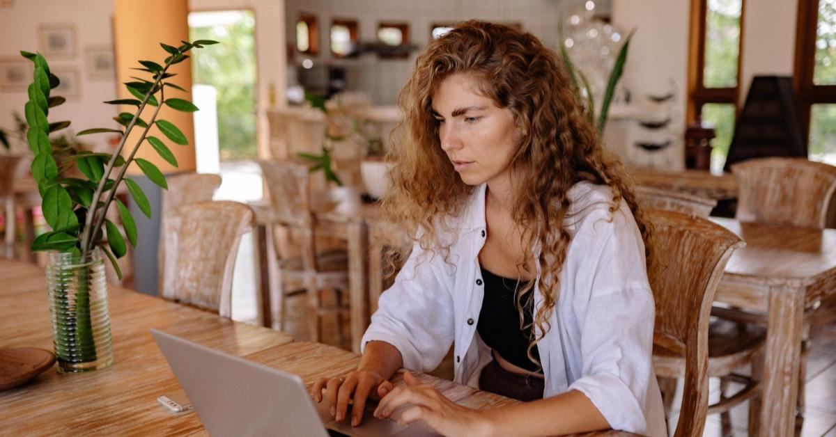 Uma mulher branca, de cabelos cacheados castanho-escuro com mechas claras, está sentada numa mesa, de frente para seu notebook. Ela usa uma camisa aberta branca, e uma blusa preta. A sala em que ela está simboliza uma casa, decorada com plantas, itens de madeira e tons luminosos. A imagem faz alusão a uma pessoa empreendedora que usa a técnica de order bump