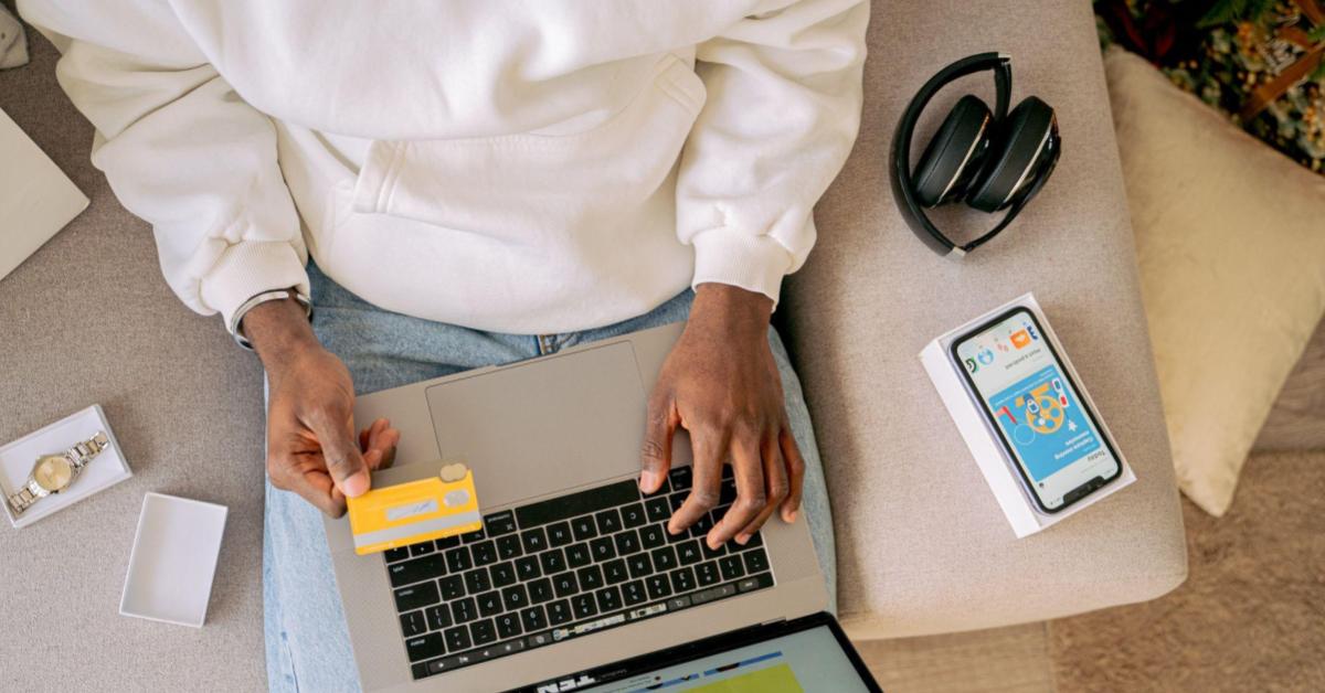 A imagem mostra um homem negro, de blusa branca e calça jeans sentado num sofá bege, enquanto busca por um produto, no site que observa em seu notebook, que está no seu colo. Ao seu lado há uma caixa branca com um rel[ogio prata, um celular e um fone, itens que dão a alusão a compras feitas no e-commerce durante datas comemorativas.