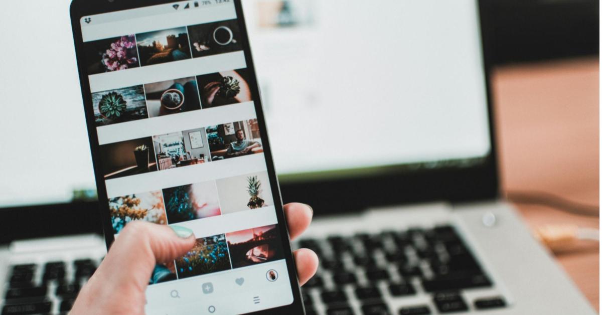 A fotografia mostra uma mão segurando um celular, em que a tela mostra a página do instagram. Ao fundo é possível ver um computador. A imagem faz menção a um planejamento que considera os melhores horários para postar nas redes sociais.