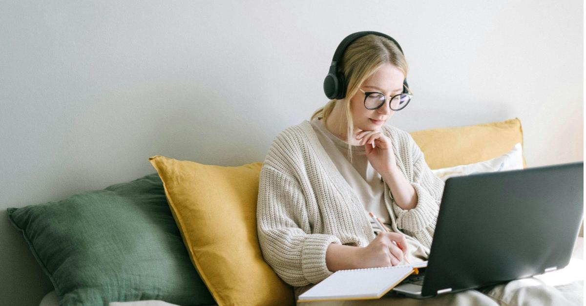 A imagem mostra uma mulher loira, branca, com os cabelos presos, sentada em um sofá com almofadas grandes, amarelas e verdes escuradas. Ela usa fones de ouvido e uma blusa de moletom off white. Enquanto está sentada, ela apoia um notebook em seu colo, dando a impressão de que está fazendo uma pesquisa, por exemplo, de um bom erp para sua loja virtual.