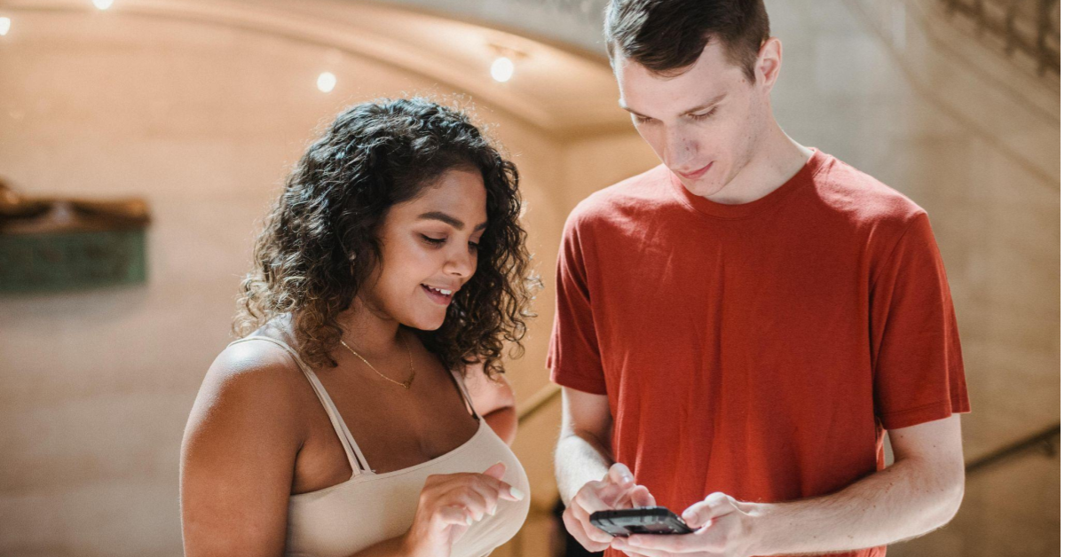a imagem mostra um homem branco, de camiseta vermelha, e uma mulher, de cabelos castanhos longos e encaracolados, vestida com uma blusa bege, mexendo em um celular, como se estivessem procurando por uma dúvida em um faq no e-commerce