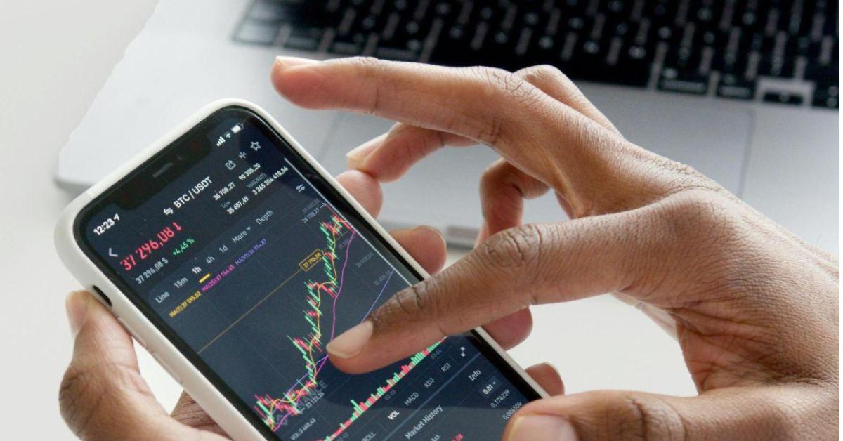 A imamgem foca na mão de uma pessoa preta, mexendo em um celular. A tela do celular mostra um aplicativo com um gráfico em crescente. Uma alusão ao momento que uma pessoa estuda taxa de importação.